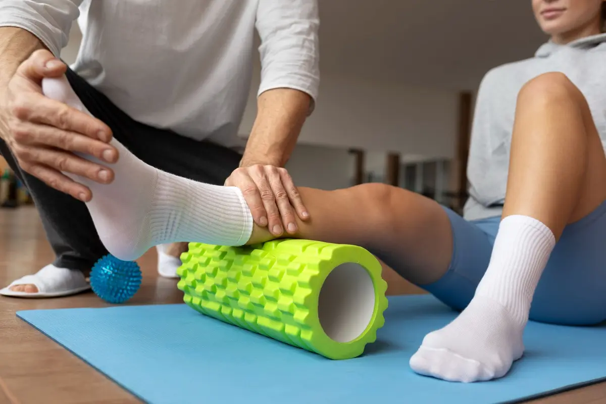 Photo of a person sitting on a yoga mat and receiving help to perform a leg exercise using a green foam roller, focusing on the calf muscle. The individual helping is wearing a white shirt and gray pants, while the person exercising is in a gray sweatshirt and blue leggings.