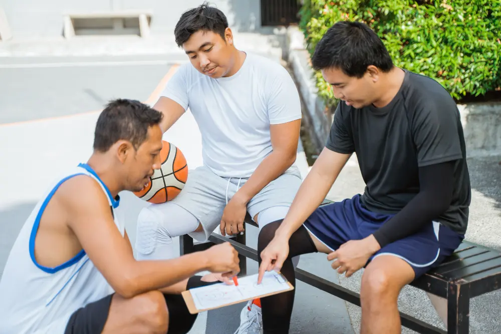 A coach in a white T-shirt and two male athletes, one in a blue tank top and the other in a black T-shirt, are discussing strategy over a clipboard on a bench beside a basketball court. The coach appears to be explaining tactics with the players attentive.
